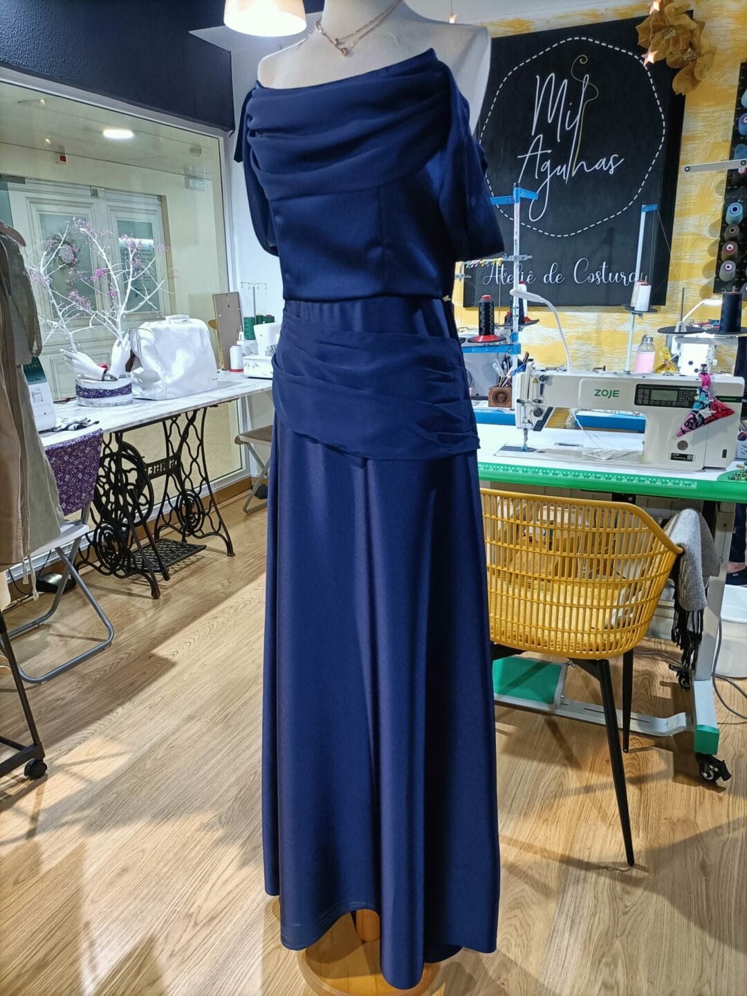 Vestido longo de festa em cetim azul escuro, com decote drapeado e franzido na cintura, exibido num manequim num ateliê de costura. Máquinas de costura e carretéis de linha visíveis ao fundo.