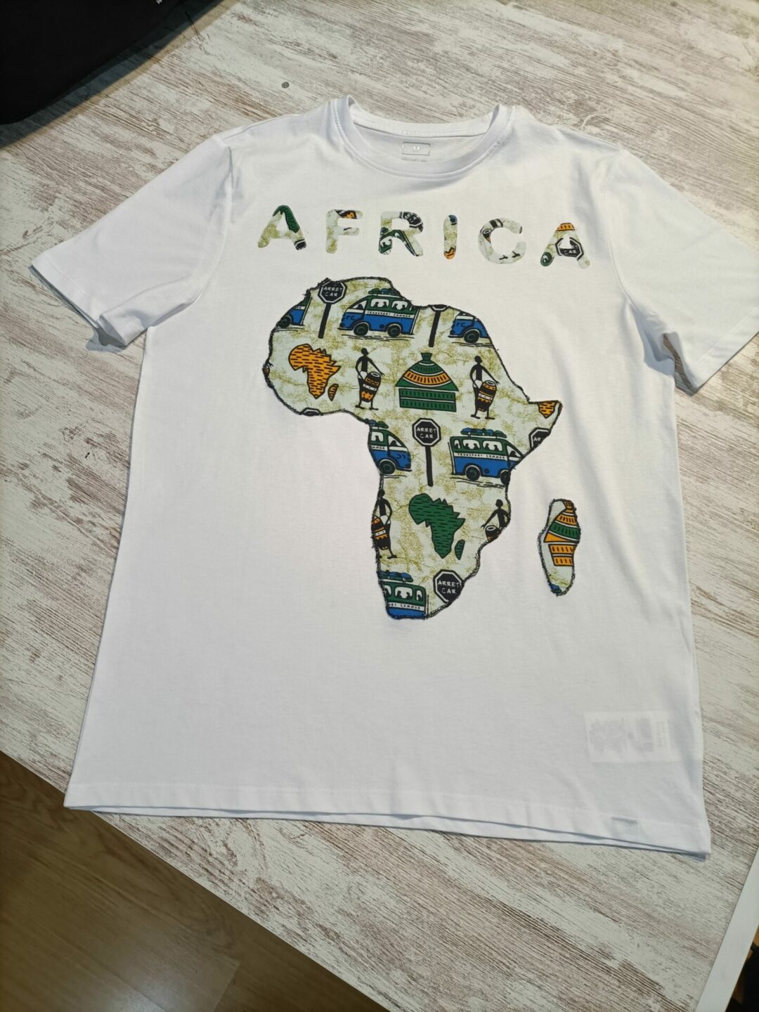 Uma t-shirt branca de manga curta deitada sobre uma superfície de madeira clara. No centro da t-shirt, um aplique em tecido estampado multicolorido representa o mapa do continente africano. Acima do mapa, a palavra "AFRICA" é formada por letras individualizadas, também em tecido estampado.