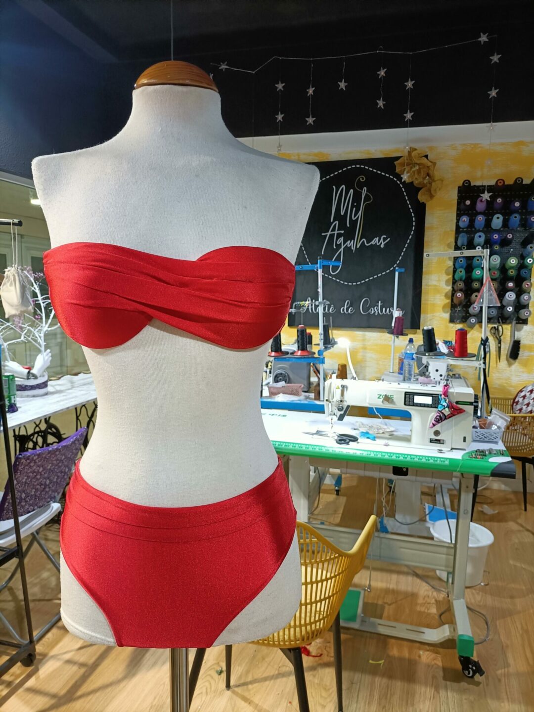 Um biquíni vermelho de duas peças (parte de cima bandeau e cueca clássica) exibido num manequim de corpo inteiro dentro de um ateliê de costura. Máquinas de costura e carretéis de linha são visíveis ao fundo.