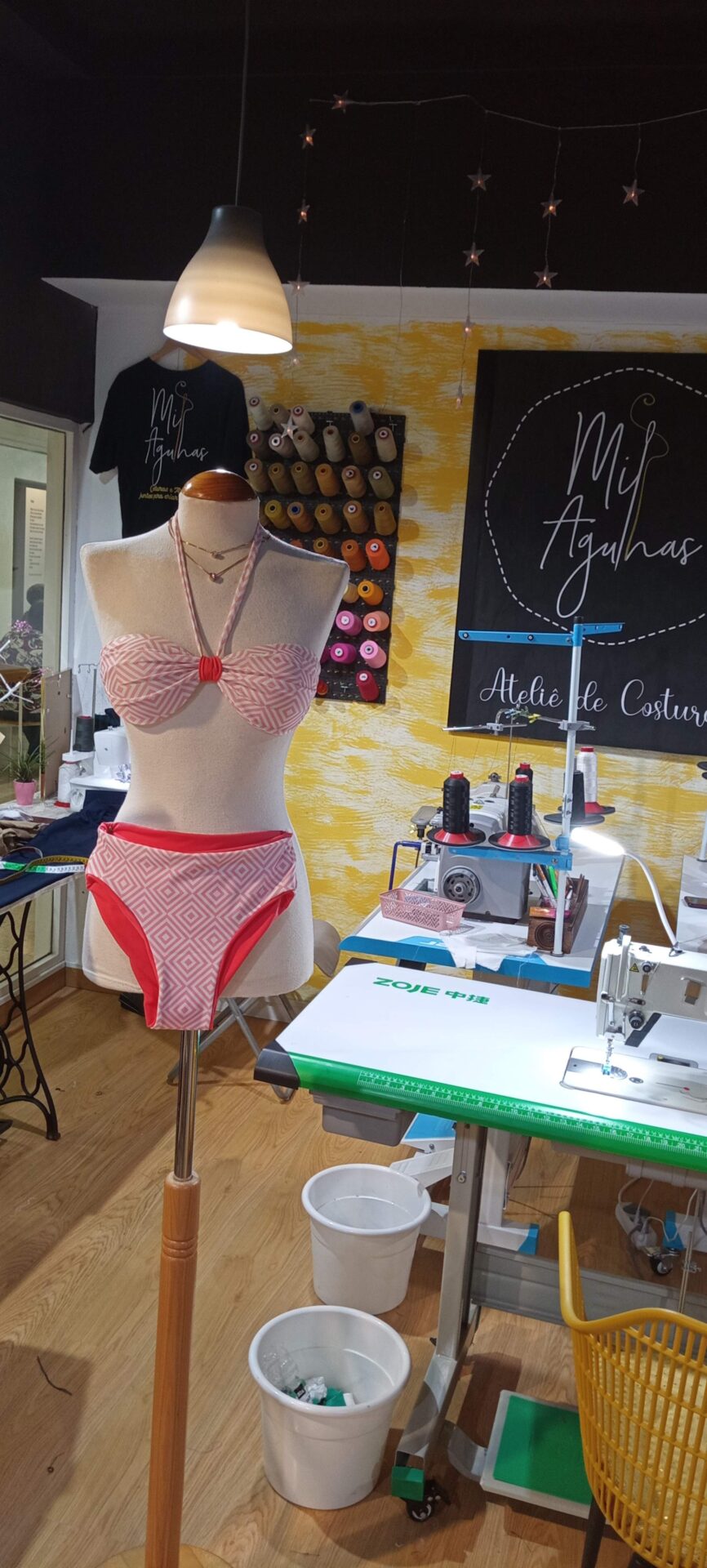 Um manequim exibe um biquíni rosa de duas peças, com a parte de cima em padrão geométrico branco e rosa e laço frontal, e a parte de baixo lisa em rosa. O manequim está dentro de um ateliê de costura com máquinas e linhas ao fundo.