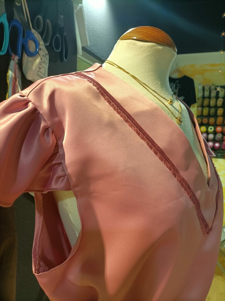 Close-up de um decote em V de uma blusa rosa em tecido acetinado ou seda, exibido num manequim. O decote possui um acabamento com uma fita decorativa em tom ligeiramente mais escuro. Duas correntes douradas finas com pequenos pendentes delicados adornam o pescoço do manequim.