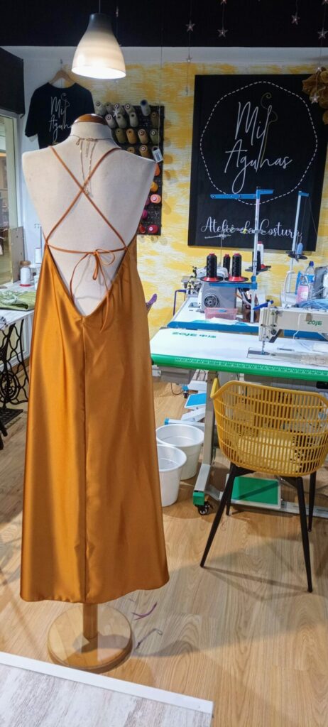 Vista das costas de um vestido longo em cetim dourado ou marigold, com alças finas que se cruzam e atam na parte inferior das costas, exibido num manequim em ateliê de costura.