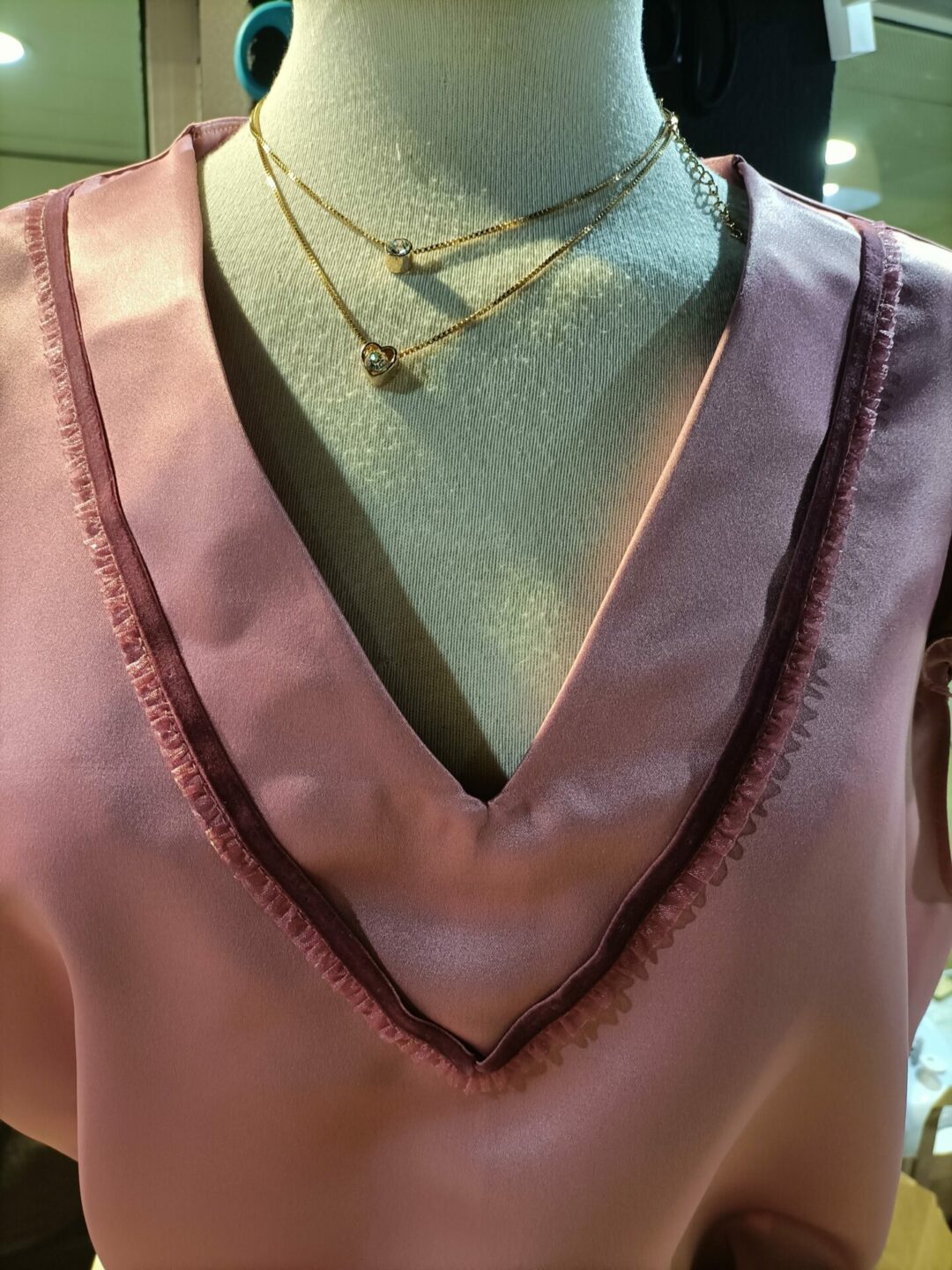 Close-up de um decote em V de uma blusa rosa em tecido acetinado ou seda, exibido num manequim. O decote possui um acabamento com uma fita decorativa em tom ligeiramente mais escuro. Duas correntes douradas finas com pequenos pendentes delicados adornam o pescoço do manequim.