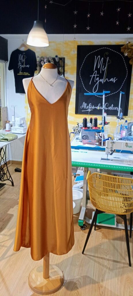 Um vestido longo de cetim/seda em tom dourado ou marigold, com alças finas e decote em V, exibido num manequim de madeira dentro de um ateliê de costura. Máquinas de costura e o logótipo "Mil Agulhas" são visíveis ao fundo.