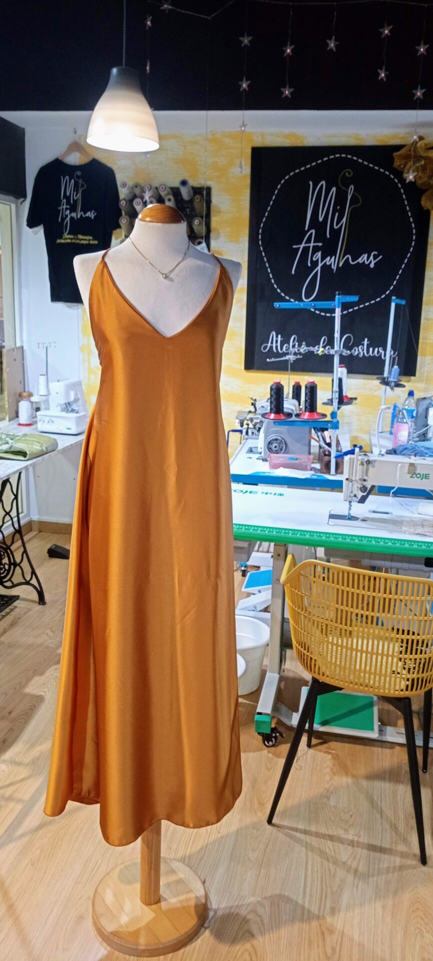 Um vestido longo de cetim/seda em tom dourado ou marigold, com alças finas e decote em V, exibido num manequim de madeira dentro de um ateliê de costura. Máquinas de costura e o logótipo "Mil Agulhas" são visíveis ao fundo.