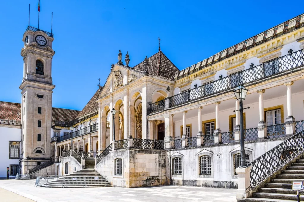 Vista panorâmica da Universidade de Coimbra, Portugal, mostrando a Torre do Relógio, o Paço das Escolas e outros edifícios históricos sob um céu azul. Há uma estátua em primeiro plano à direita. A imagem simboliza a riqueza cultural da cidade visitada por um estudante Erasmus.