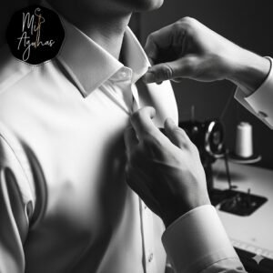 Um homem a abotoar a manga de uma camisa branca, enquanto uma costureira, à direita, segura e ajusta o colarinho. O foco está nos detalhes do tecido e das mãos a trabalhar na camisa. O logótipo "Mil Agulhas" está visível no canto superior esquerdo. A foto está a preto e branco para um efeito clássico.