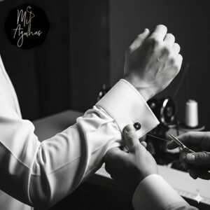 Close-up das mãos de um alfaiate ou costureiro a ajustar o punho de uma camisa branca de homem, com uma tesoura e linha de costura visíveis, sugerindo reparação ou confeção. A imagem está a preto e branco e inclui o logótipo "Mil Agulhas" no canto superior esquerdo.