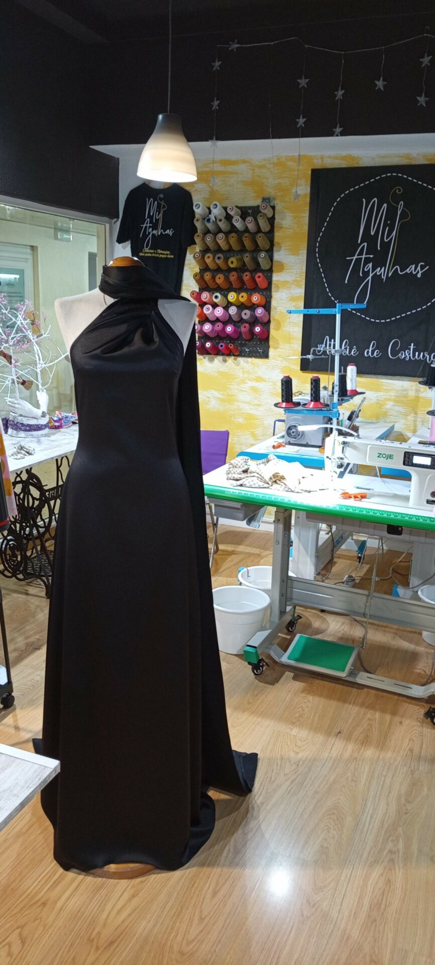 Vestido de alta costura preto em cetim, com gola alta e capa fluida num ombro, exposto num manequim no ateliê Mil Agulhas.