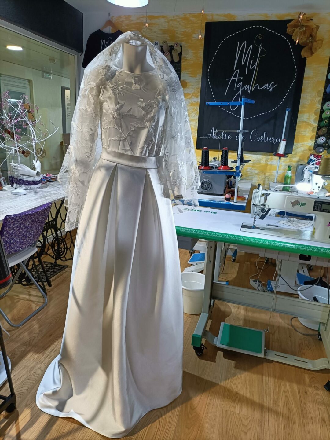 Vista frontal de vestido de noiva Mil Agulhas com saia de cetim acetinado, corpo de renda floral com pérolas, mangas compridas e véu alto, no ateliê em Coimbra.