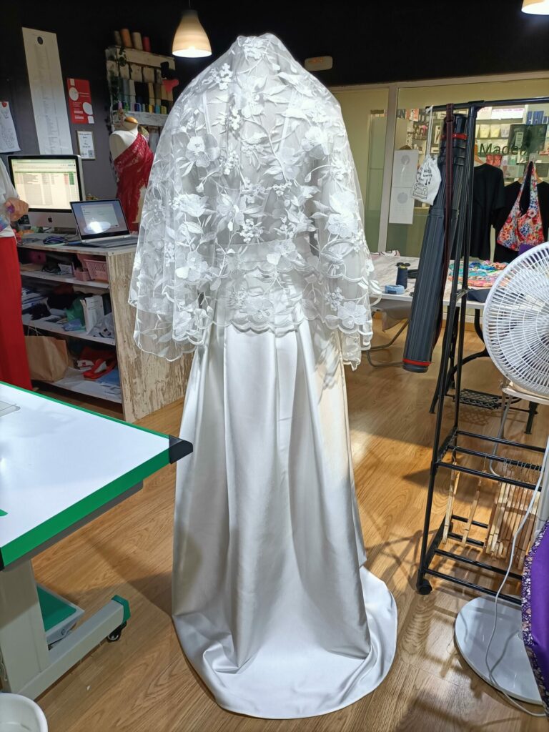 Vista de costas de vestido de noiva Mil Agulhas, destacando o véu artesanal em renda floral que cobre os ombros e a cauda discreta da saia de cetim.