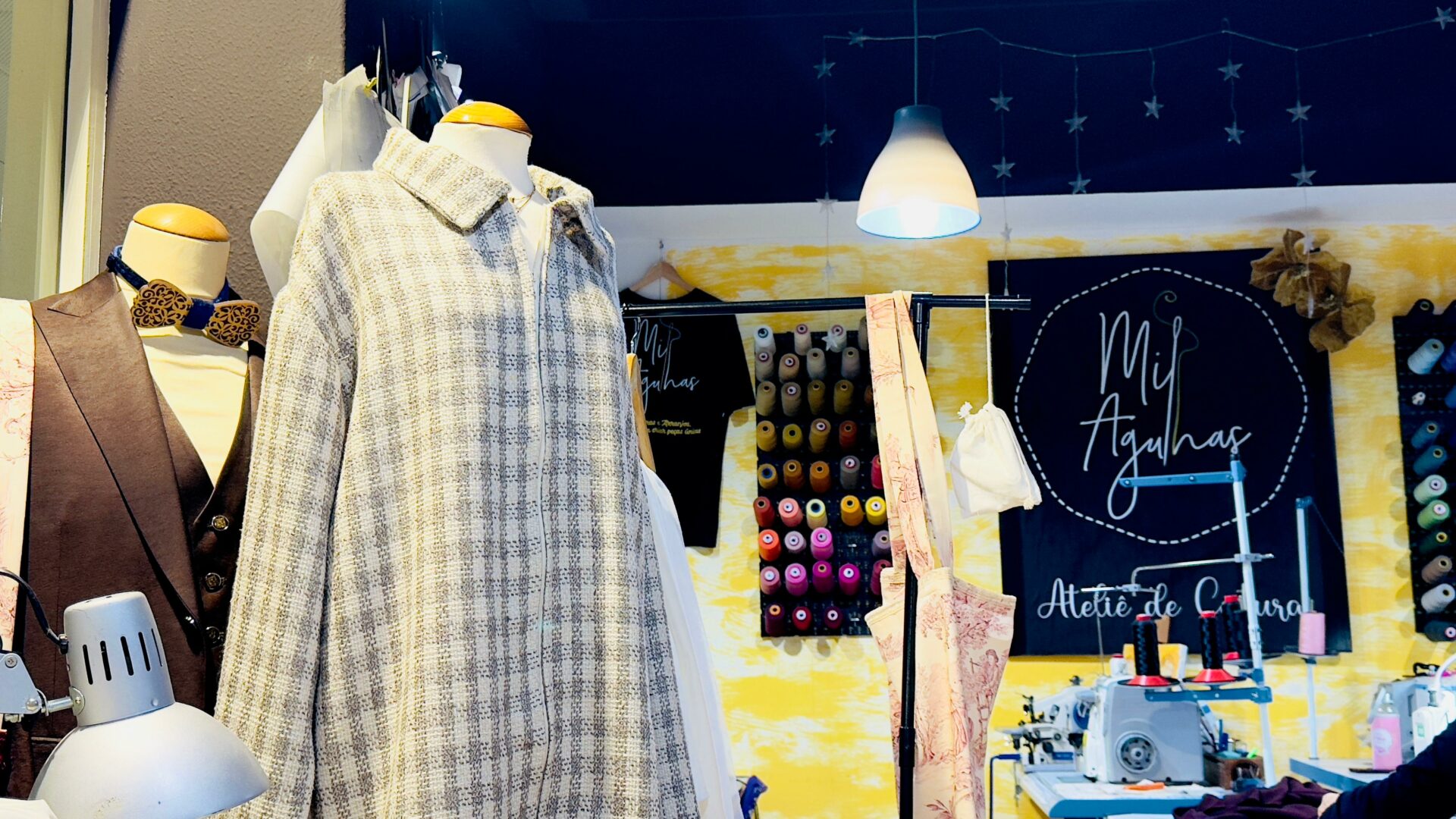 Ateliê de Costura “Mil Agulhas” e Criações em Exibição Manequins num ateliê de costura. Um manequim em primeiro plano veste um camisão xadrez cinza e branco, e ao fundo vê-se uma máquina de costura e um painel com carretéis de linha coloridos.