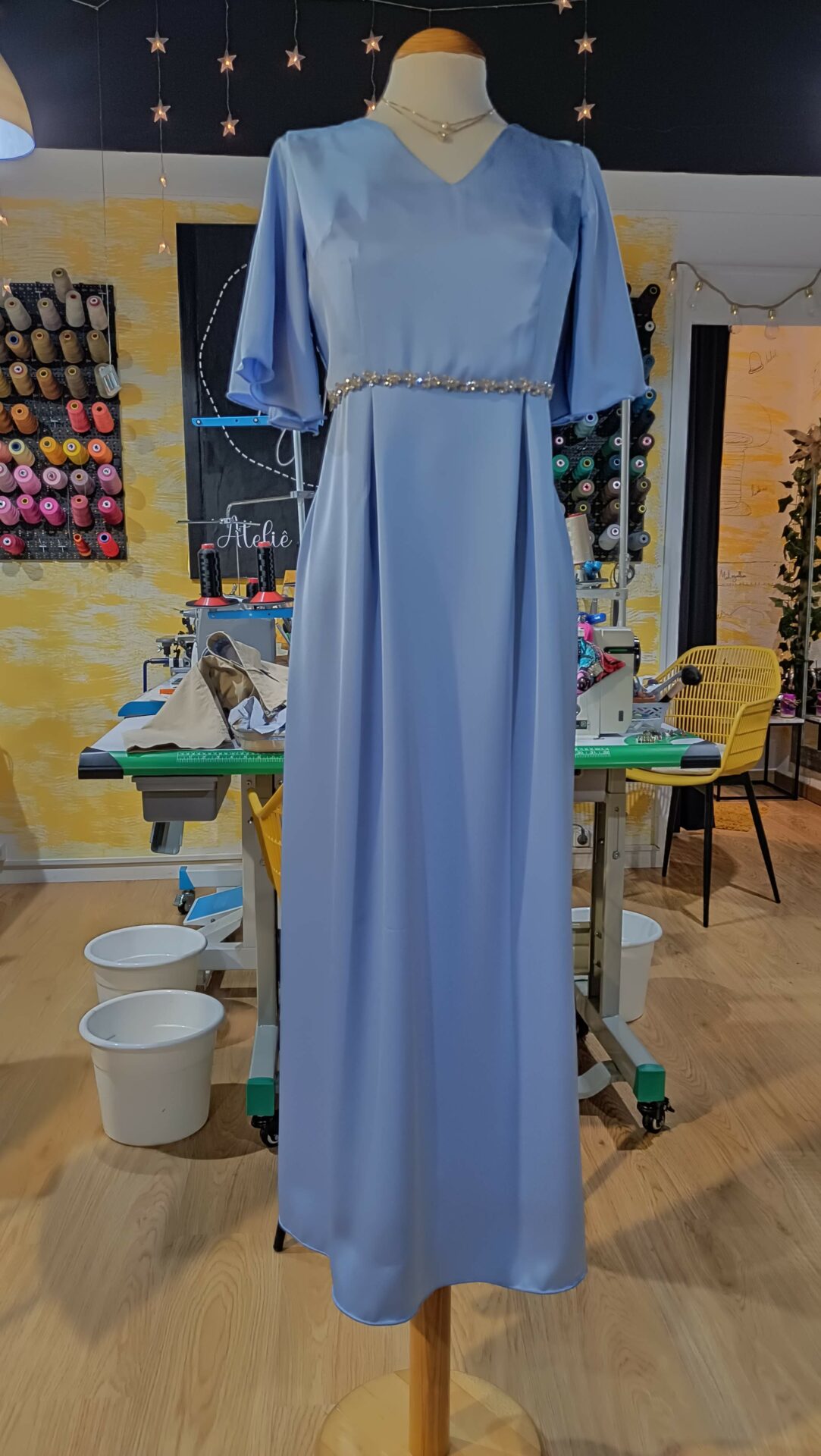 vestido-cerimonia-azul-serenidade-mil-agulhas-coimbra Vestido de cerimónia comprido, em azul claro, com mangas esvoaçantes e cinto de brilhantes, exposto num manequim no ateliê de costura Mil Agulhas em Coimbra.