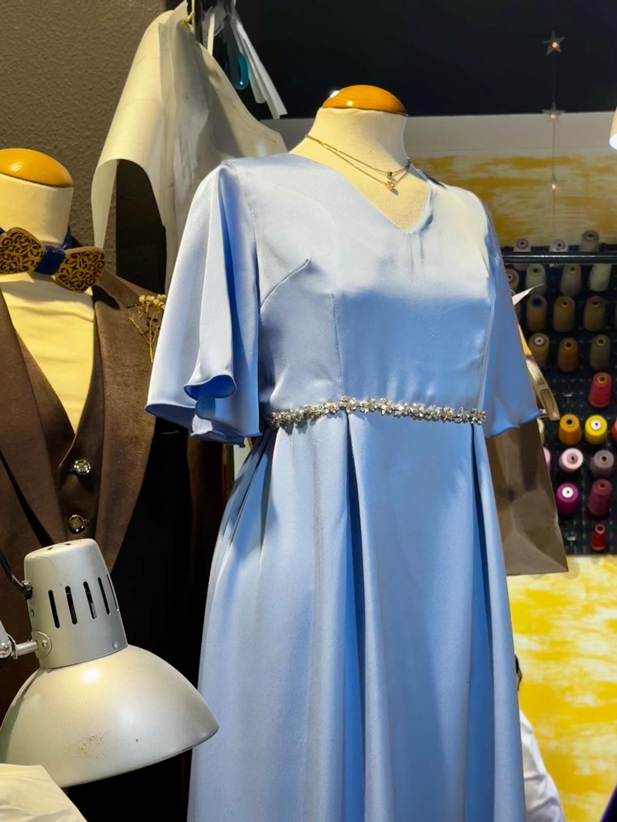 vestidocerimoniaazulserenidade-mil-agulhas-coimbra Vestido de cerimónia azul claro com mangas esvoaçantes e cinto de brilhantes num manequim, com um manequim masculino de colete castanho e laço dourado ao lado, dentro do ateliê de costura.