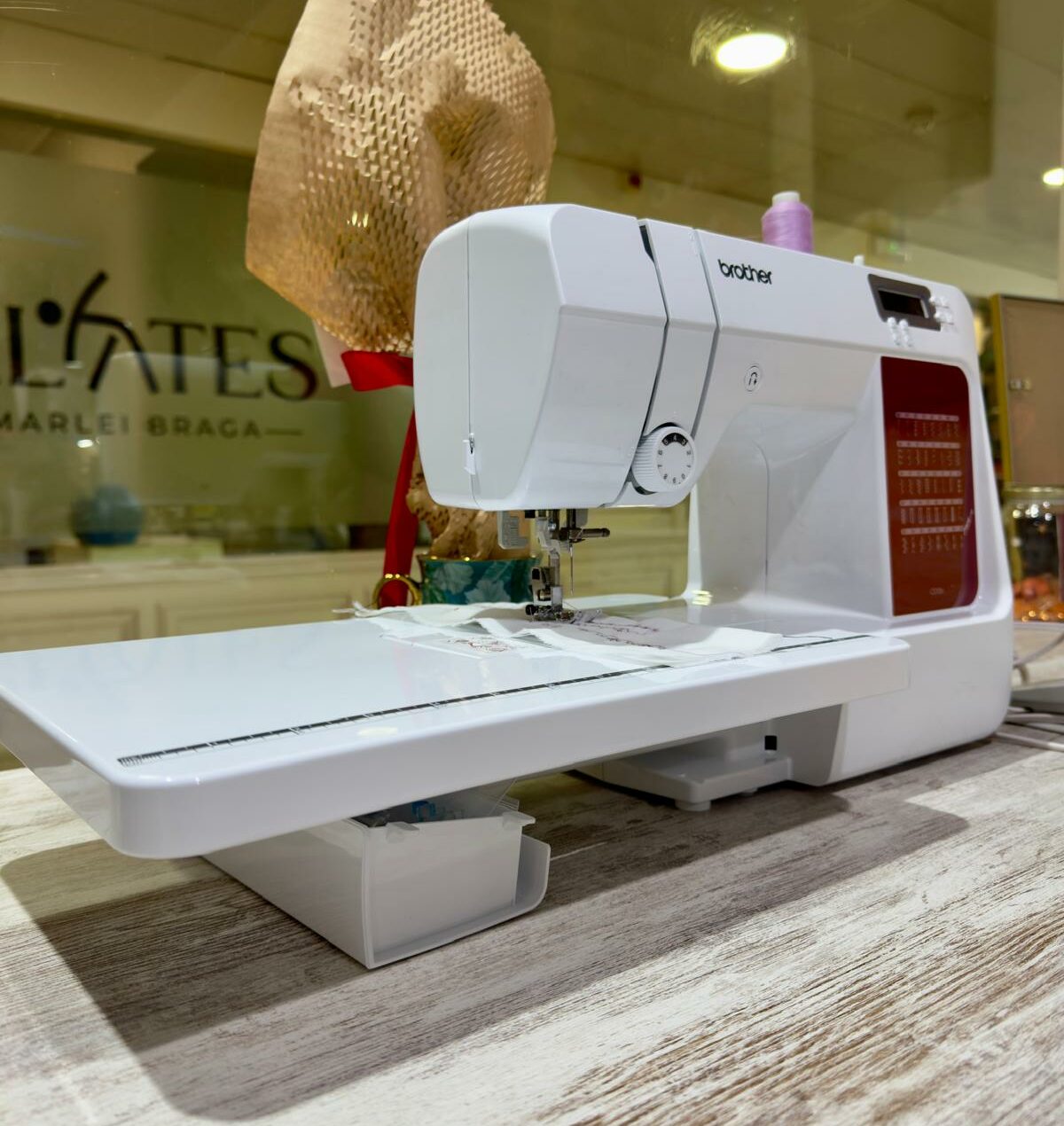Máquina de costura Brother branca com mesa de extensão e tecido bordado em primeiro plano, num atelier moderno.