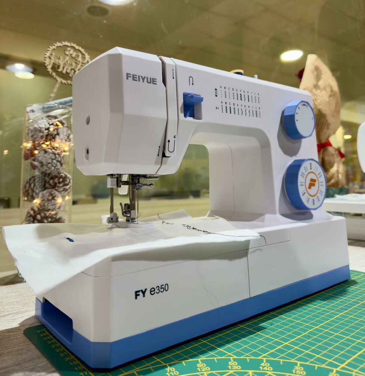 Máquina de costura Feiyue FY e350 branca e azul com tecido e pinhas decorativas, num ateliê de costura.