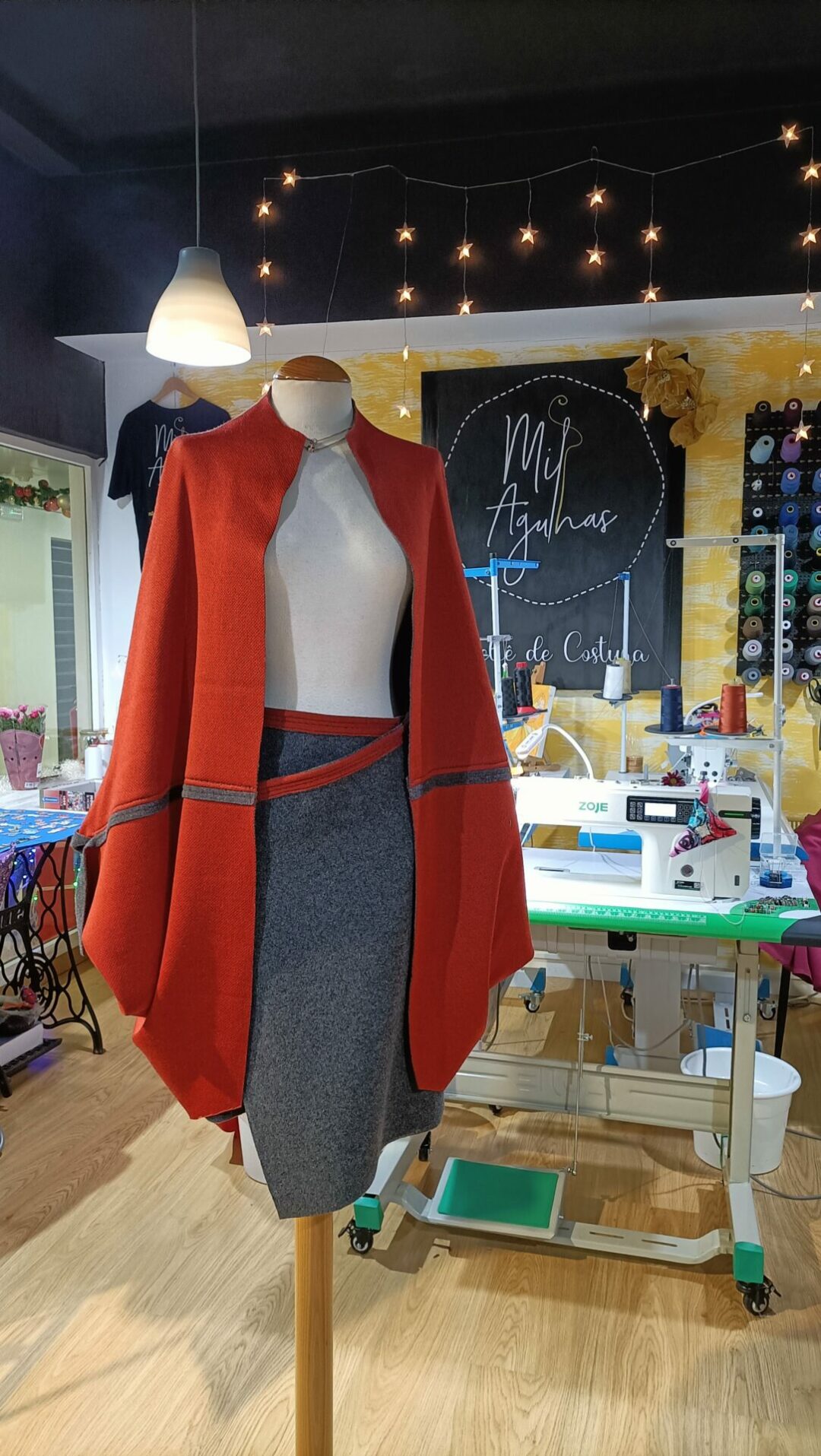 Conjunto feminino composto por uma capa estruturada vermelha com debrum cinza e saia lápis cinza mesclado, exibido num manequim dentro do ateliê Mil Agulhas.