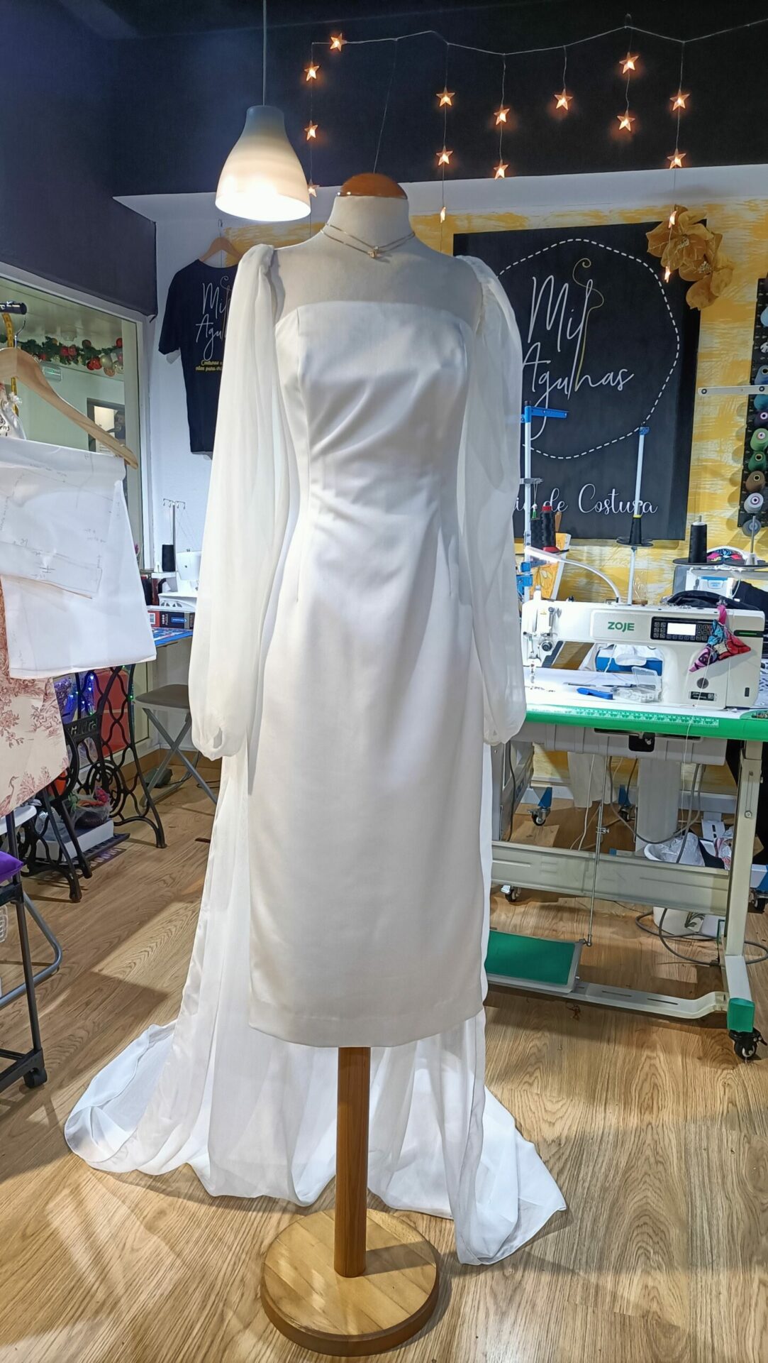 Vista frontal de um vestido de noiva minimalista branco em manequim, com decote quadrado, corpo estruturado e mangas bufantes em organza transparente num atelier de costura.
