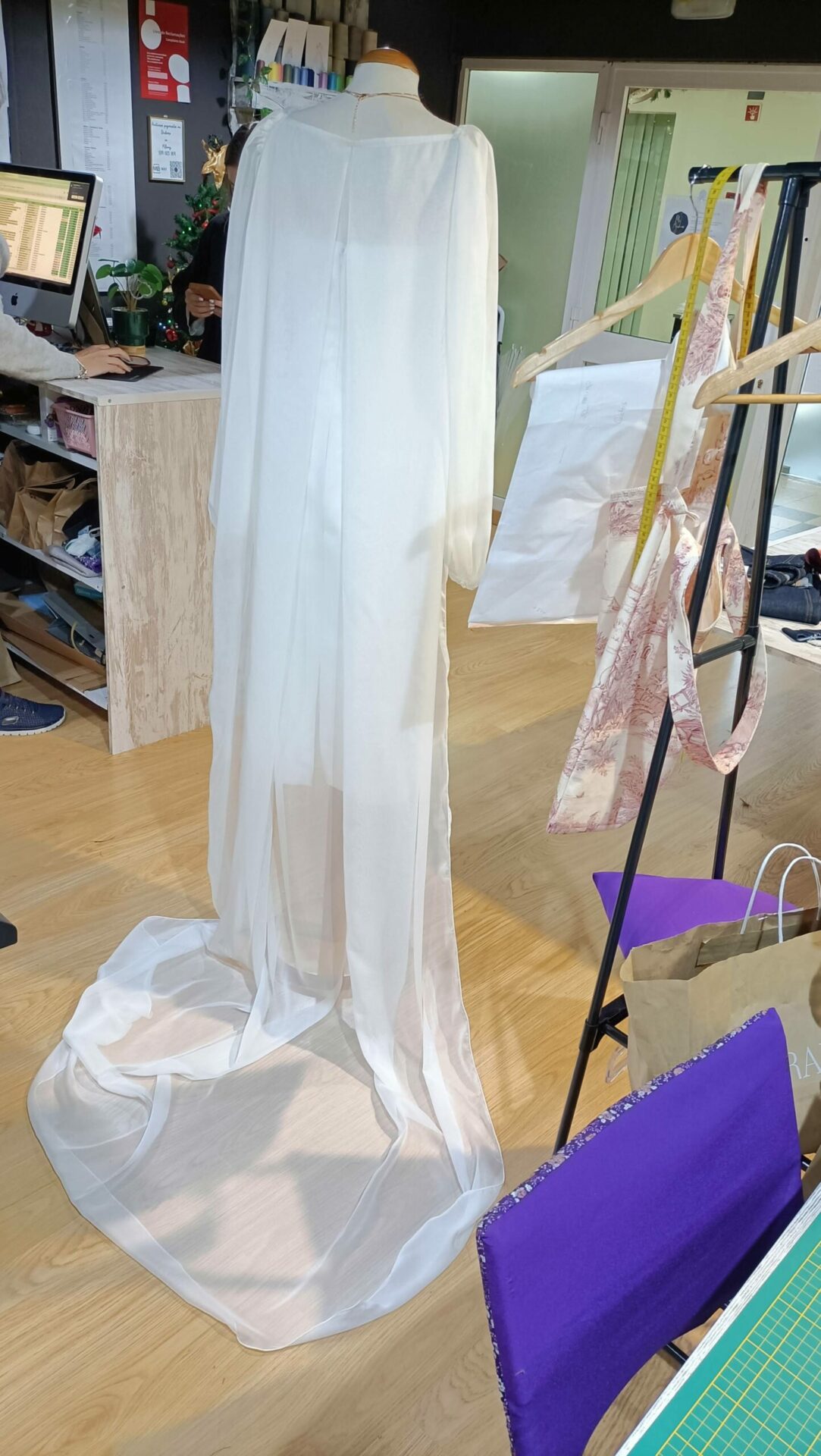 Vista traseira de um vestido de cerimónia branco com uma capa fluida de organza que se estende numa cauda elegante sobre o chão de madeira do atelier Mil Agulhas.