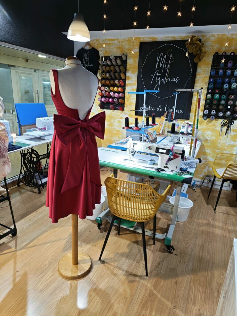 Vista traseira de um elegante vestido de cerimónia vermelho feito à medida, destacando as costas descobertas e um grande laço na cintura. O vestido está exposto num manequim dentro do ateliê de costura Mil Agulhas, rodeado por máquinas de costura e linhas.