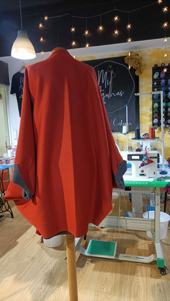 Vista de costas de uma capa feminina vermelha de corte amplo e fluido, destacando o caimento do tecido e os detalhes cinza nos punhos.